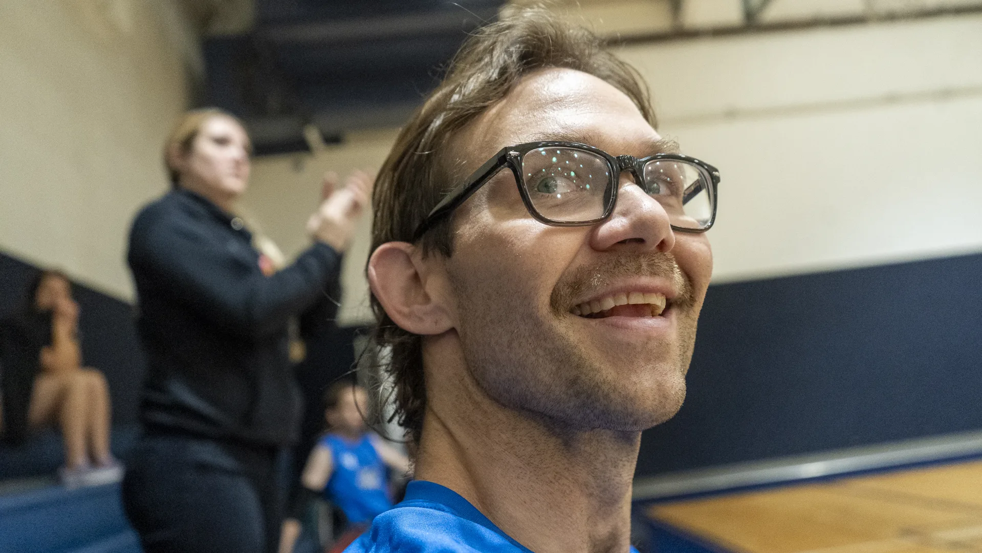 A smiling man wearing dark-framed glasses and a blue shirt looks upward in a gym.