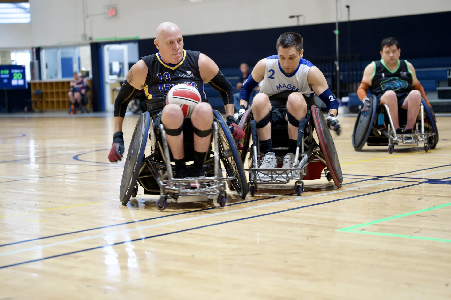Three wheelchair rugby players race across an indoor court, with the leading player carrying a red-and-white ball on his lap.
